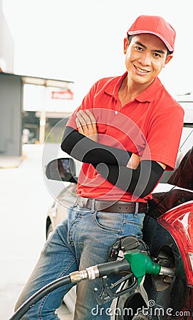 Asian gas station worker man leaning against black car and looking straight with smile while green fuel nozzle filling high energy Stock Photo