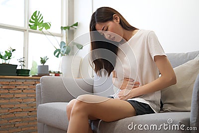 Asian girl having period is sitting on sofa and feeling much of painful on her stomach with unknown reason. She holds onto her Stock Photo