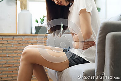 Asian girl having period is sitting on sofa and feeling much of painful on her stomach which called women`s pain or menstrual Stock Photo