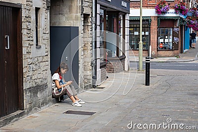 An Asian lady in her twenties looking at her mobile phone whilst sat on a step outside a shop Editorial Stock Photo