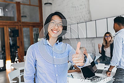 Asian laughing boy posing with thumb up at the beginning of the workday. Chinese office worker in blue shirt and glasses Stock Photo