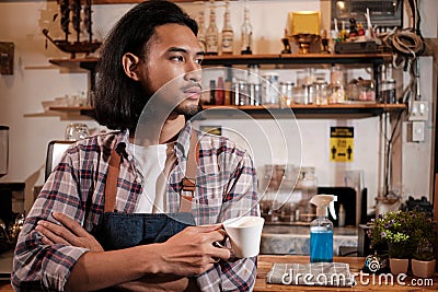 Asian male barista stands at a casual cafe, looking outside with a coffee cup Stock Photo