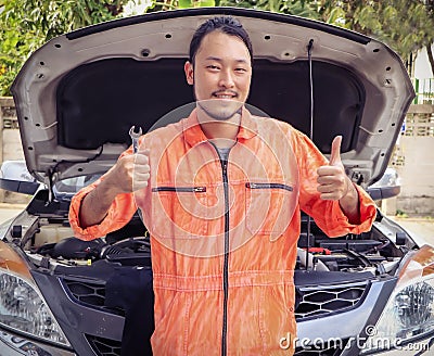 Asian Male mechanic wearing orange color jumpsuit working on car engine outdoor, standing in front of car holding wrench and Stock Photo