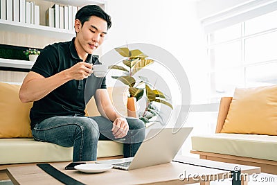 Asian man sitting and working on the sofa while drinking coffee. Businessman in casual working on laptop at home. Stock Photo