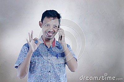 Asian man talking on the phone over isolated grey background doing ok sign with fingers, smiling positive doing ok sign with hand Stock Photo