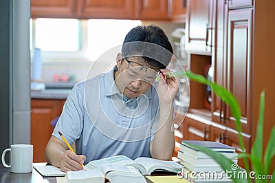 Asian middle-aged man sitting at desk at home, reading a book and studying Stock Photo
