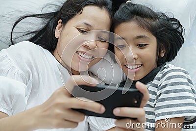 Asian mother and daughter playing game with phone together on the bed. Women and girl happy and fun in bedroom Stock Photo