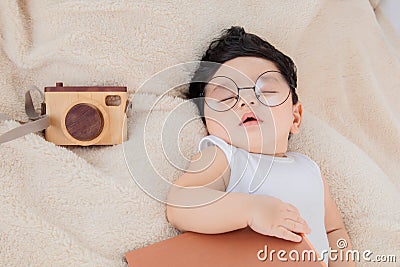 Asian Newborn baby wearing glasses with favorite book on beige blanket , 3 month-old infant lying in bed with relax. Adorable baby Stock Photo