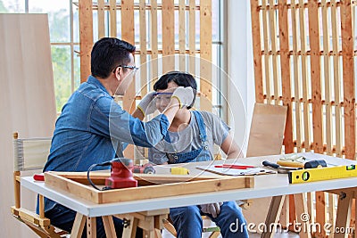 Asian professional carpenter engineer architect woodworker dad in jeans outfit with safety goggles gloves touching young boy son Stock Photo
