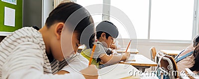 Asian school children sitting at desk in school writing in note book with pencil, studying, education, learning. Male Stock Photo