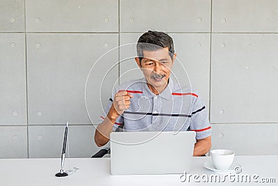 Asian senior man. with casual clothes. Rejoice, raise fist right hand. Sitting look at to laptop computer screen. Stock Photo