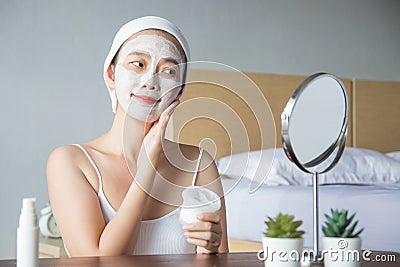 Asian woman applying moisturizer face scrub peeling white clay mask on skin, looking in mirror with smiley face using cream Stock Photo