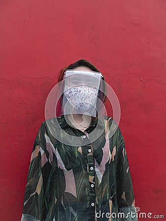 An asian woman with a face mask and face shield looks at a camera. Agaisnt a red wall. Stock Photo