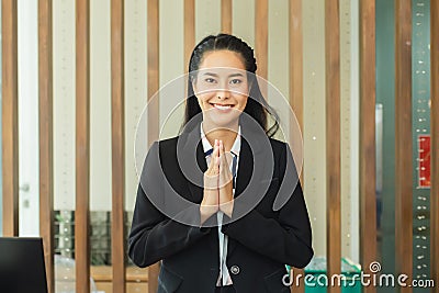 Asian woman hotel receptionist smiling and hand up wai thai style greetings hand to pay respect standing at front of lobby. Stock Photo