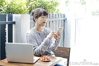 Asian woman sitting at a table and using smartphone at home. Typing text message, via cell phone, Social networking concept. Stock Photo