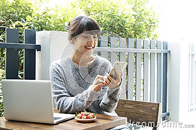 Asian woman sitting at a table and using smartphone at home. Typing text message, via cell phone, Social networking concept. Stock Photo