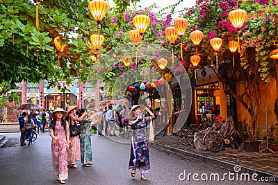 Hoi An, Vietnam. Asian dressed women stroll through an avenue in the tourist attraction, in whose trees hang illuminated lanterns Editorial Stock Photo