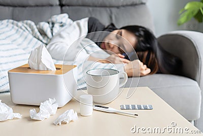 Asian woman suffering from covid-19 symptoms have to be on self-isolation at home. Girl has a fever lying on couch with tissue, Stock Photo