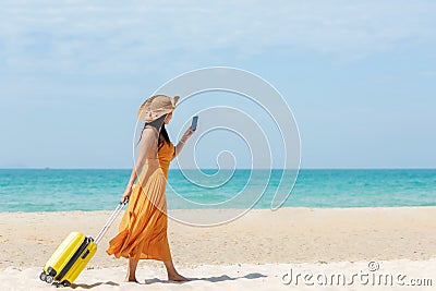 Asian women traveler holding yellow luggage walking and check in smart phone on the beach Stock Photo