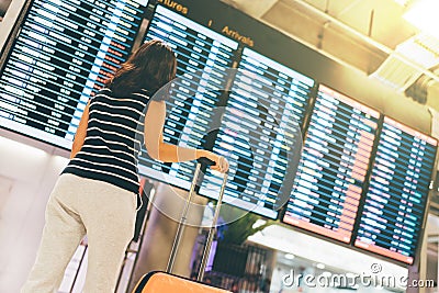 Asian woman traveler looking at flight information screen in an airport, holding suitcase, travel or time concept Stock Photo