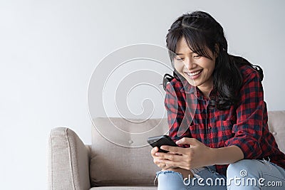 Asian woman are using a smartphone while on sitting sofa in the living room at home. Woman wearing a red plaid shirt look Stock Photo