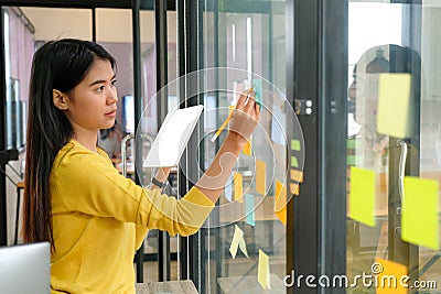 Asian woman wearing a yellow shirt with a tablet in her hand She is writing a pencil on a note on a glass wall in the office Stock Photo