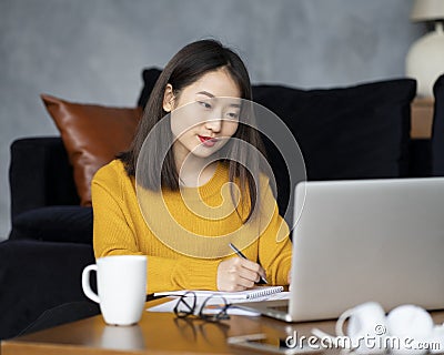 Asian woman working on laptop at home or in hotel. Young lady in bright yellow jumper Stock Photo