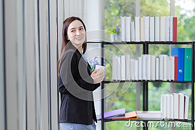 Asian working lady in a black suit holds paper cup to drink coffee in the morning looking out of the office window Stock Photo