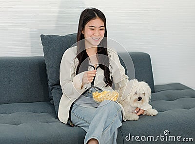 Asian young happy cheerful female owner sitting smiling on cozy sofa couch holding eating popcorn glass bowl snack watching movie Stock Photo