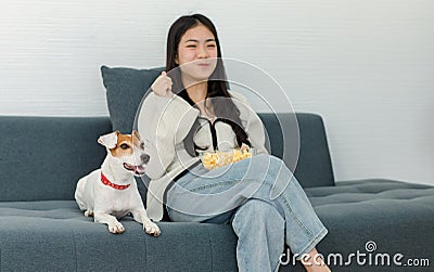 Asian young happy cheerful female owner sitting smiling on cozy sofa couch holding eating popcorn glass bowl snack watching movie Stock Photo