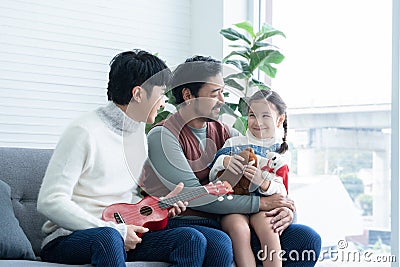 Asian young LGBTQ gay couple playing ukulele and singing with little Caucasian adopted kid in living room at home Stock Photo