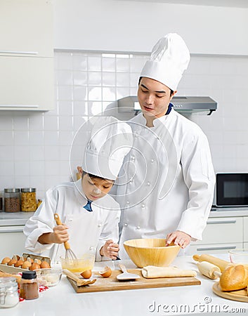 Asian young little boy pastry chef in white uniform with tall cook hat standing while male cooking teacher smiling help teaching Stock Photo