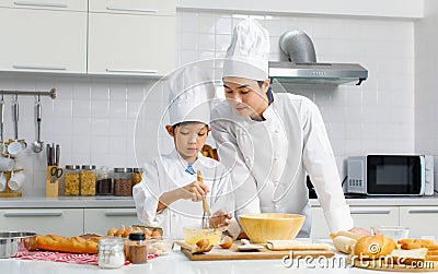 Asian young little boy pastry chef in white uniform with tall cook hat standing while male cooking teacher smiling help teaching Stock Photo