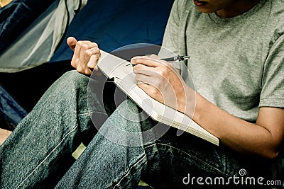 Asian young man sitting is holding a pen writing note of letter memorize memories on book in outside the tent. Loneliness Stock Photo