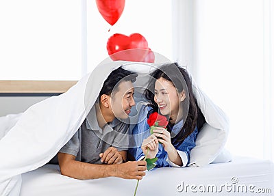 Asian young romantic lover couple male boyfriend giving red rose surprising female girlfriend laying down under blanket on bed Stock Photo