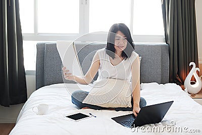 Asian young woman is sitting on white bed and reading papersheet for online working and learning with laptop in social medea in Stock Photo