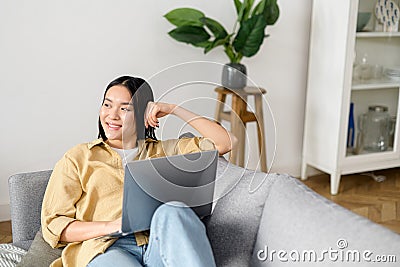 Asian young woman spending home leisure with laptop. Calm girl sitting at cozy sofa and looking away in front of the Stock Photo