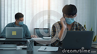 Asians sit in their office and use the phone to talk to clients while wearing masks in their offices during COVID-19 Stock Photo