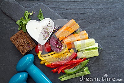 Assorted fresh chopped vegetables with dip and dumbbells on dark background, sport and healthy food concept Stock Photo