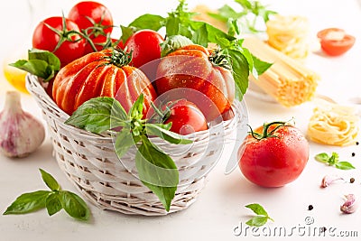 Assorted tomatoes with basil in basket, garlic, spice and raw pasta for italian cuisine. Healthy food concept on white background Stock Photo