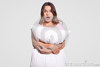 Astonished dark haired woman carries white soft pillow, horrified by nightmare, surprised by something terrible, keeps jaw dropped Stock Photo