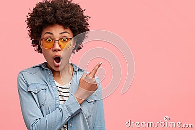 Astonished fearful black young woman drops jaw with fright, keeps eyes popped out, points with index finger at upper Stock Photo
