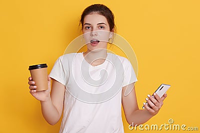 Astonished lady with opened mouth standing against yellow background, holding smart phone and take away coffee in hands, wearing Stock Photo