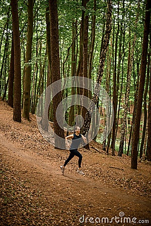 Athlete young woman in black sportswear jogging downhill on forest trail, tall leafy trees on background Stock Photo