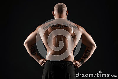 Athletic bald, bearded, tattooed man in black shorts is posing against a black background. Close-up portrait. Stock Photo