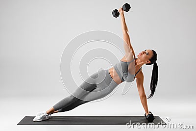 Athletic girl doing side plank exercise on gray background. Fitness woman working out with dumbbells Stock Photo