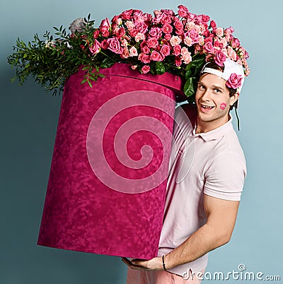Smiling man delivery guy with rose at ear and lipstick kiss on his cheek is holding huge valentines day box with flowers Stock Photo