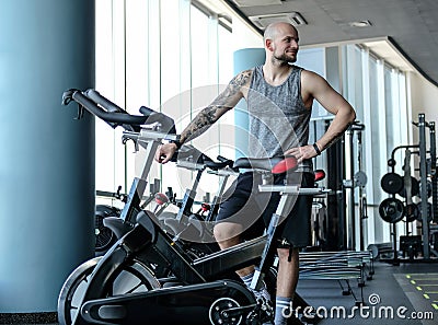 Portrait of a bald athletic man with a tattoo on his hand standing next to a exercise bike in the modern fitness club Stock Photo
