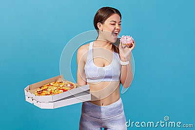 Athletic slim woman in white sportswear losing willpower and biting sweet junk donut, making choice holding in hand pizza box, Stock Photo