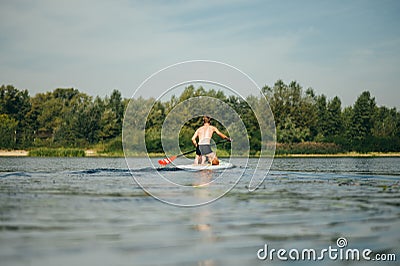 Athletic young man swims along the river on a paddle board, view from the back. Athlete trains on a sup board, actively paddles Stock Photo